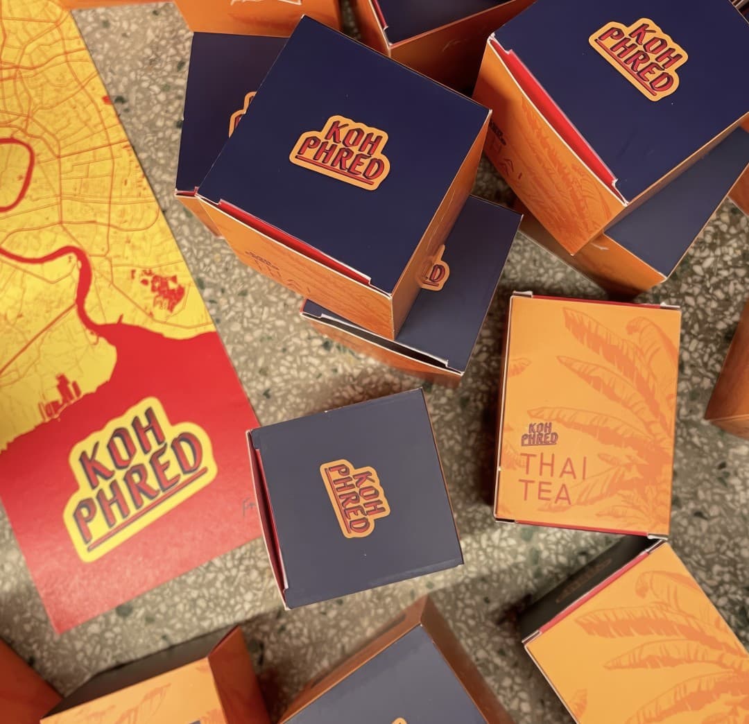 A stack of orange and navy Koh Phred packaging boxes, one labelled Thai Tea